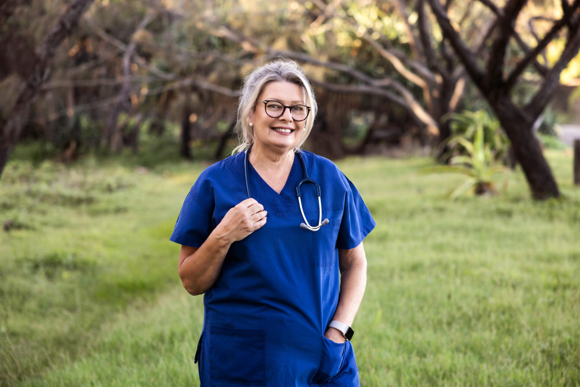 Woman in blue scrubs with stethoscope around neck, holding it with one hand and other hand in pocket, smiling
