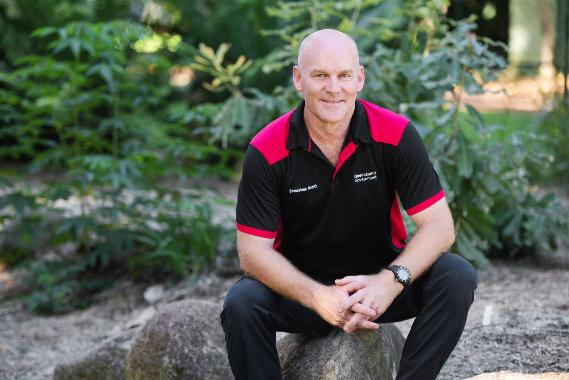 Man in Queensland Government polo shirt sat on a rock, clasping hands together and smiling