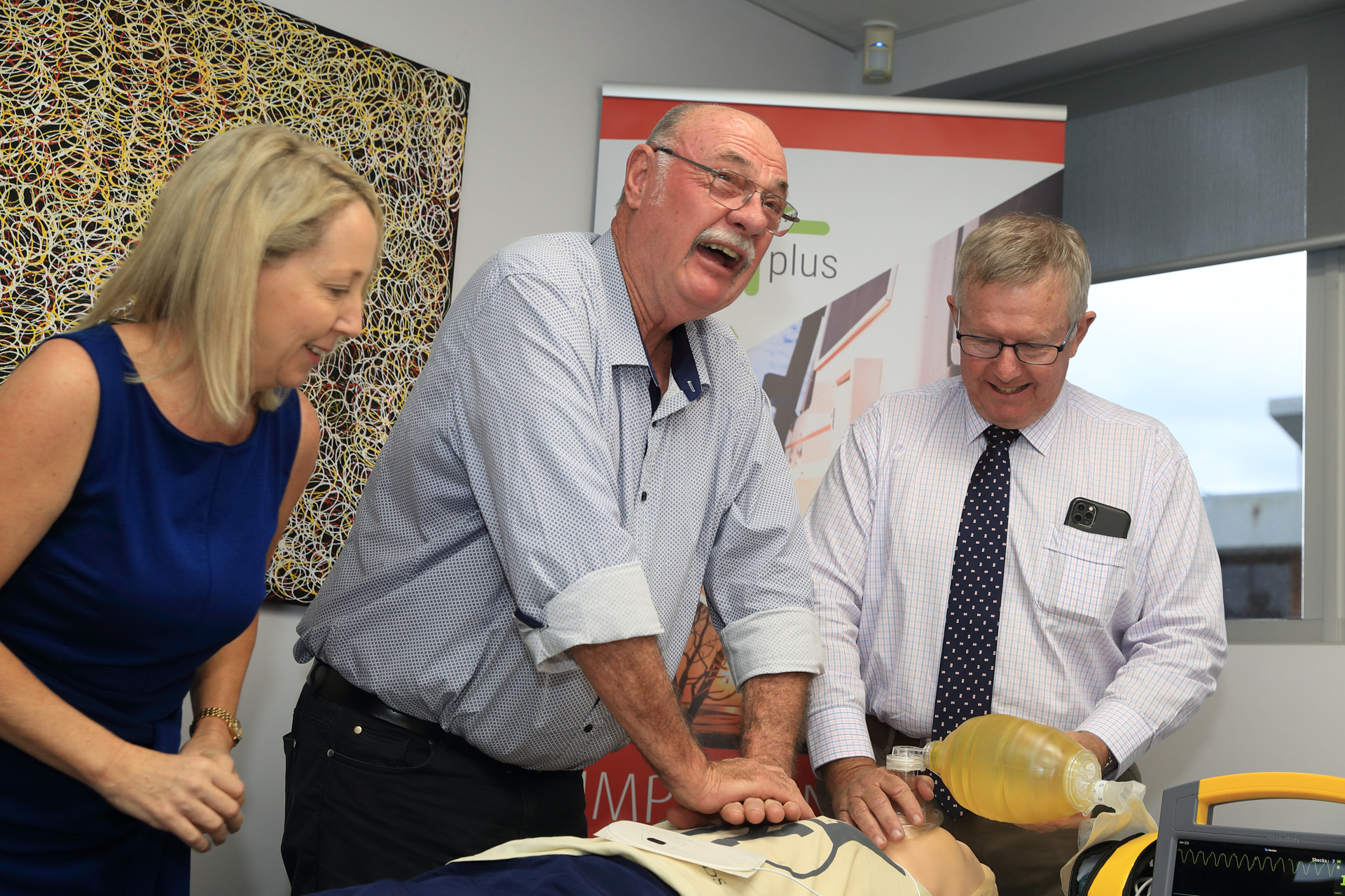 FRFP5551 Man in shirt performs CPR on a mannequin. Another man in shirt holds air mask over mannequin's mouth. Woman in dress watches.